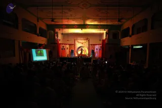 A dimly lit indoor hall viewed from the back, with rows of seated audience members silhouetted in the foreground. On the raised stage at the far end, a lone performer in dark clothing stands before a large circular golden backdrop with a glowing lamp, flanked by red and orange drapes and figures of painted panels. A projection screen on the left shows a green-toned image. The ceiling is ornately painted with floral motifs and hanging lights, and the walls feature arched windows. The scene suggests a satsang or evening programme in progress. No speaker is clearly identifiable as a person from this distance.