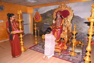 A photograph inside an orange-walled hall with golden lamp stands and a golden lion-armed throne. A seated figure in red-and-orange robes with long swept-back hair, a red crown-like headdress, a rudraksha mala and ceremonial gold necklaces raises a hand in blessing on an ornate golden throne with a red cushion, in front of a mountain temple backdrop. a person by these cues. To the left, a woman in a red sari with gold jewellery holds a brass plate of flowers and petals. A young girl in a pink lace shawl kneels on the carpet before the throne, offering flowers at the foot of the seat. Brass oil lamps flank the scene.