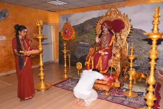 A photograph in an orange-walled room. A seated figure on an ornate gold throne with carved lion armrests raises a hand in blessing; wearing maroon and gold robes, a red turban, a rudraksha mala and a long gold necklace, and holding a staff. A woman in a maroon sari with a long braid stands at left holding a brass tray with flowers and a lit lamp. A man in white kneels at the foot of the throne, touching flowers placed on the carpet. Tall brass oil lamps stand on both sides, and a backdrop shows a temple gopuram with mountains. Swamiji (probable) is seated on the throne, identifiable by the maroon and gold robes, turban, rudraksha mala and staff.
