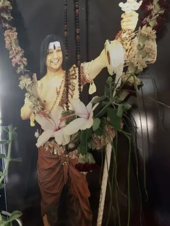 A large printed portrait of a bare-chested man with long dark hair and a dark forehead mark, wearing rudraksha mala necklaces and a rust-brown dhoti, stands against a dark background. He holds a raised ceremonial vessel with a flower-decorated top, and garlands of pink and white flowers hang around the frame and across his arm. Pink lilies and greenery are arranged at the figure's waist and along the frame. The image is a photograph of a printed portrait, so no living people are present in the scene.