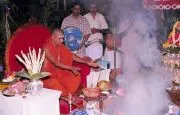 A photograph of a fire ritual at night. A young man in saffron-orange robes with long swept-back dark hair sits cross-legged before a smoking fire pit in front of a deity murti, performing a homa with offerings laid out on a brass tray and a kalasha. Behind him two men in white dhotis stand in the background among smoke, and a decorated shrine with flower garlands is visible at right. Only the priest and two standing attendants are clearly visible.