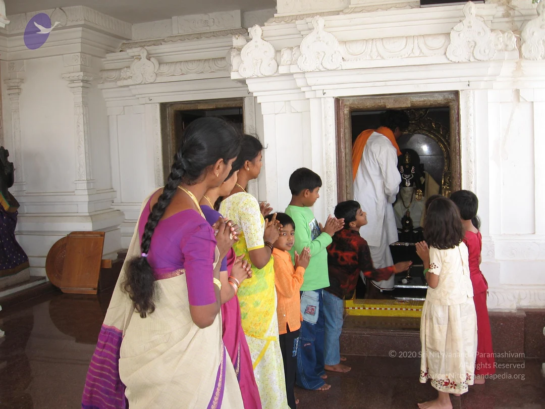 A photograph inside a white stone temple shrine showing a line of worshippers with palms pressed together in prayer before a deity murti in a dark niche. A woman in a cream saree with a magenta blouse and a long braid stands at left, followed by a woman in a yellow and white outfit, then several children in green, orange, red-brown and blue clothing and a girl in a cream dress at right. Inside the shrine a man in white dhoti with an orange shawl stands before the murti, which is adorned with garlands and a gold necklace. The floor is polished dark stone and the walls carry carved white ornamentation.