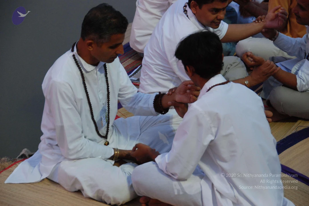 A photograph of seated participants in an indoor meditation therapy session on a mat-covered floor. In the foreground, a man in white kurta and white trousers, wearing a dark wooden rudraksha mala and a bangle on his wrist, sits cross-legged while holding hands with a younger man in white who sits with his back partly turned. Behind them, more seated people in white and light-coloured clothing hold hands in a circle, with a grey wall in the background. The scene suggests a group energy or healing exercise.