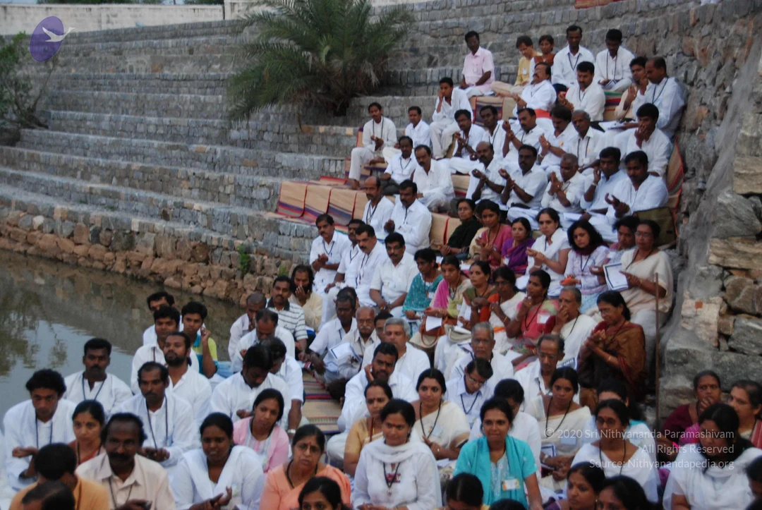 A large outdoor gathering of devotees seated on stone steps beside a water tank at dusk. Dozens of people, mostly dressed in white with blue lanyard badges, sit on striped mats in rows, many with hands pressed together in namaskar. Some women wear saffron, pink, teal and turquoise saris and salwar, and a few carry papers. A small palm tree grows on the upper terrace, and the stone wall rises behind the crowd. No a person candidate is visible among the seated participants.