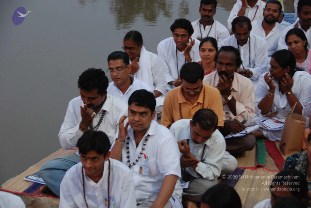 A group of seated devotees in white clothing sits on mats beside a still water body. Many wear rudraksha malas around their necks. Several men in white shirts and white dhotis, plus women in white shawls and saris, sit close together and rest their fingers at their lips or temples, appearing to be in meditation or a guided practice. A man in the front row wears a bindi and white shirt with a mala, and a man in an orange polo with a mala sits in the centre. Papers and a bag lie on the mats. No one is identifiable as a person in this frame.