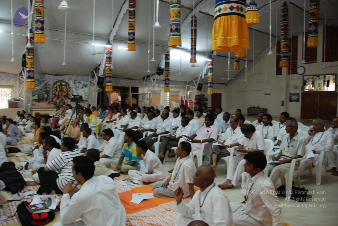A large indoor hall with white chairs and a seated audience of mostly men in white shirts and white trousers, some sitting on mats and others on white plastic chairs, several holding papers and booklets. Hanging from the ceiling are long cylindrical banners in bright red, yellow, blue and green patterns with yellow fabric drapes. At the far left stands a framed deity image on an altar near a painted backdrop. A video camera on a tripod stands at left, and a wall clock hangs on the right. A man in the front row sits with hands folded in a prayer gesture. No speaker or a person candidate is clearly visible in the frame.