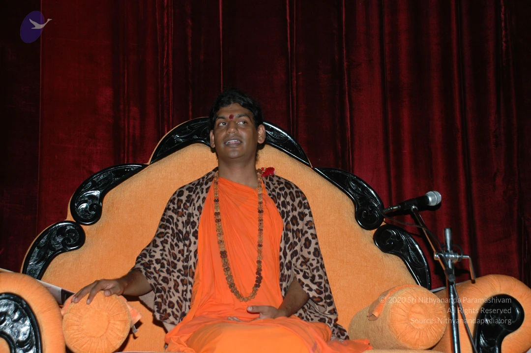 A seated man in an orange saffron robe sits on an ornate orange-upholstered throne with black carved wooden armrests and backrest, in front of deep red velvet drapes. He wears a long rudraksha-style bead mala, a leopard-print shawl draped over his shoulders, and a red bindi on his forehead, with a red flower tucked near his shoulder. He rests one hand on an orange bolster and gestures with the other while speaking into a microphone on a stand at right. a person: the seated speaker's appearance shows saffron robes, a long mala, a leopard-print shawl and a throne, which are consistent with the a person description, though the face and identity cannot be confirmed.