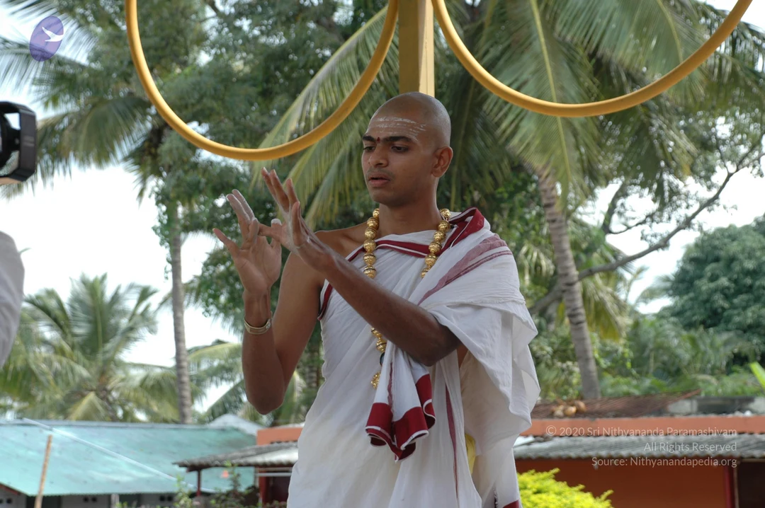 Outdoor photo at an ashram setting with coconut palms and a low orange-walled building behind. A shaved-headed man in white ritual cloth with a red-bordered shawl stands under a golden ring-shaped frame, hands raised in a mudra-like gesture. He wears a large gold bead necklace and a gold bangle, with white sacred ash markings on his forehead and body. A camera is visible at the left edge.