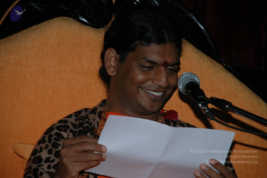 A smiling man with short dark hair and a red kumkum bindi on his forehead sits in front of an orange upholstered chair with a dark wooden frame. He wears a leopard-print shawl over orange clothing and a red flower garland at his neck. He holds an open white sheet of paper in both hands, reading it, and a silver microphone on a stand is positioned beside his face. The scene looks like a satsang or talk. No other people are visible.