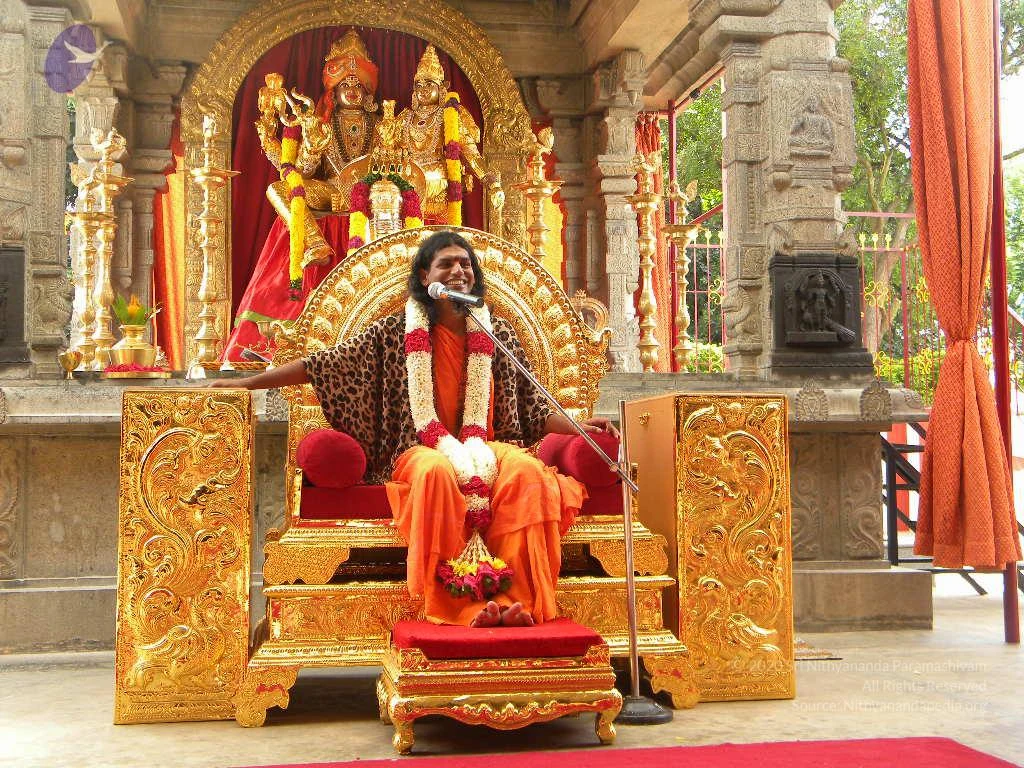 A seated man sits on an ornate gold throne with red cushions in front of a temple-style shrine with gold lamps and a red-draped niche holding deity murtis garlanded with yellow and red flowers. He wears a leopard-print shawl over orange robes, a white and red flower garland, and smiles while speaking into a microphone on a stand. Orange curtains hang at the right, and a red carpet covers the floor. A gold footstool with red cushion and a flower offering rest before him. The man has long dark hair and is barefoot, which fits the a person cues of long hair, saffron clothing and garlands on a throne.