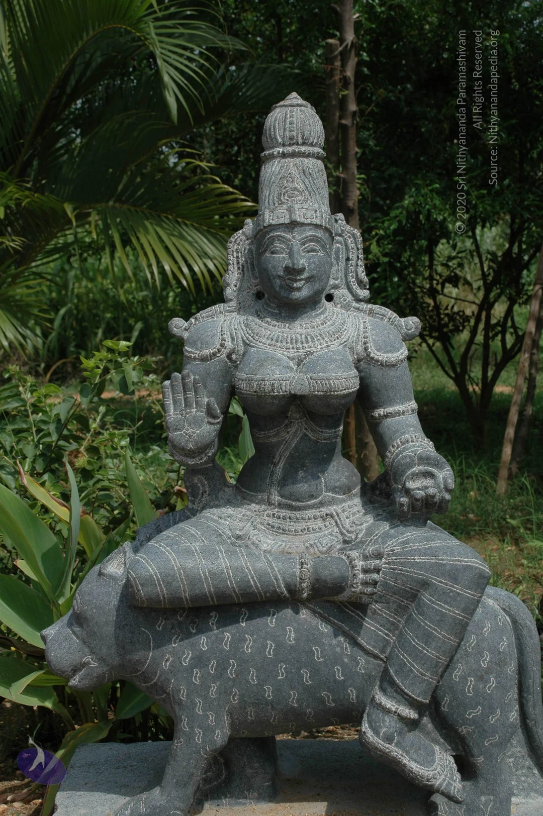 A grey stone statue of a seated deity mounted on a recumbent bull, set outdoors among palm fronds and green foliage. The figure wears a tall ornate crown, a broad necklace, armlets and bangles, and holds one hand raised in abhaya mudra while the other rests on the bull's back. The bull is carved with a pattern of small paisley-like marks. No living people are visible. Deity murti in stone, not a person; no a person candidate present.