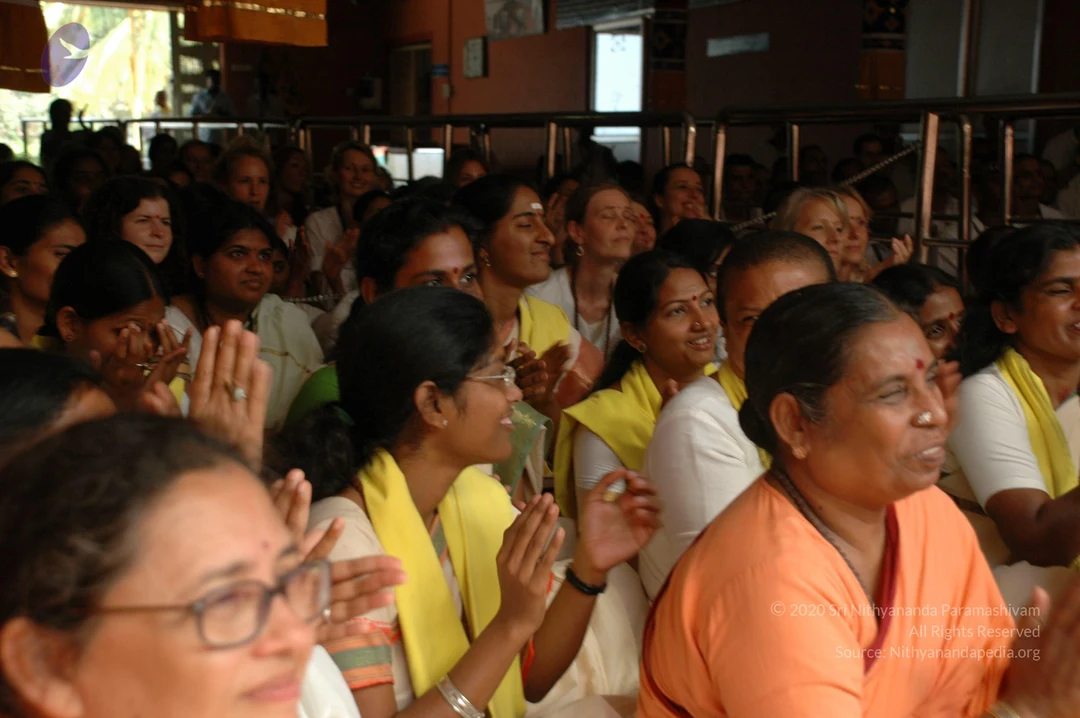 A photograph of a seated crowd of devotees, mostly women, at an indoor ashram hall with metal railings behind them. Many wear white clothes with yellow shawls or scarves, and several have their palms pressed together in greeting or prayer. A woman in the foreground wears an orange sari, a red bindi and a nose ring, and another in the lower left wears glasses. Some people are smiling and clapping. Behind the railings, more devotees and attendees are visible near open windows and a doorway.