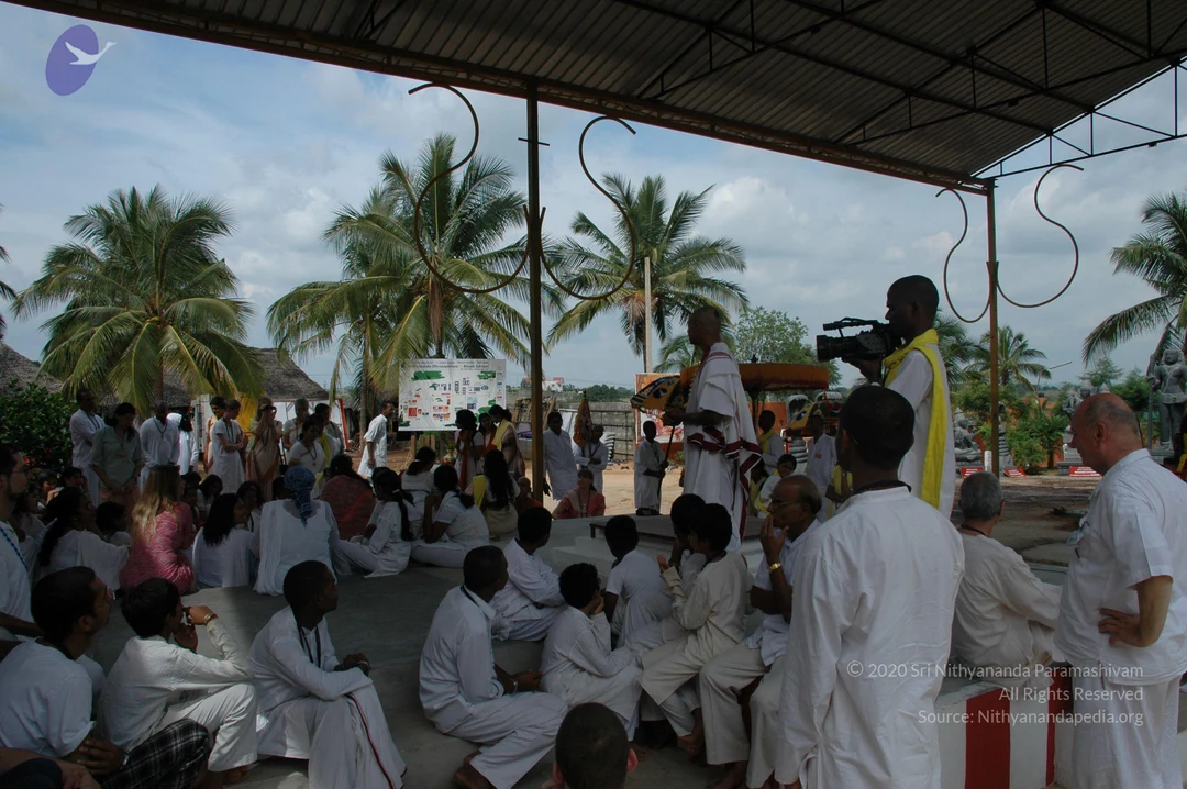 Outdoor ashram gathering under a corrugated metal roof with curved iron rods, with coconut palms and thatched huts behind. Many people in white clothes sit on a raised platform. A bald man in a white dhoti and shawl stands near a wooden stand, while a cameraman in white with a yellow shawl films him with a handheld video camera. A man in white and a bald man in white stand at the right edge. A printed chart on an easel stands in the background, and a statue is visible at the far right.