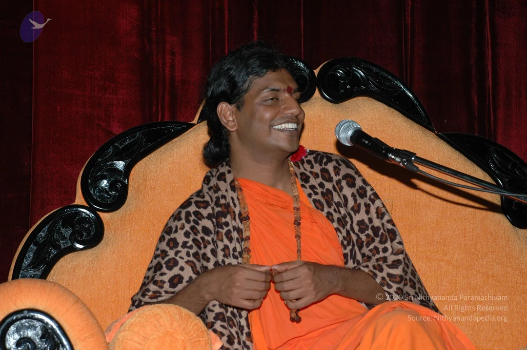 A photograph of a smiling man seated on an ornate orange velvet throne with carved black wooden armrests, in front of a deep red velvet curtain. He wears saffron-orange robes, a leopard-print shawl draped over his shoulders, a rudraksha mala around his neck, and a red bindi on his forehead. His hands are clasped in his lap, and a microphone on a stand is beside him. a person cues: long swept-back hair, saffron clothing, rudraksha mala and a throne. The image is an archive photo from an ashram event.