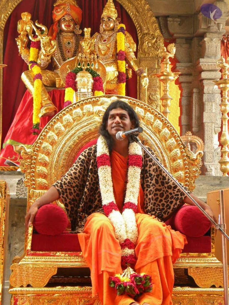 A seated man in saffron-orange robes sits on an ornate gilded throne with red cushions, a microphone on a stand in front of him. He wears a leopard-print shawl over his shoulders and garlands of white and red flowers draped across his chest and lap, with a small bouquet of red and white flowers resting on his knees. Behind him stand gold-coloured deity murtis, one wearing an orange turban and holding a trident, decorated with yellow and red flower garlands. Gold lamp stands and carved stone pillars frame the scene. Looking at the central figure, the saffron robes, long swept-back hair, and ceremonial garlands are appearance cues consistent with the page's claim, so he is described as a person seated for a satsang-style address.
