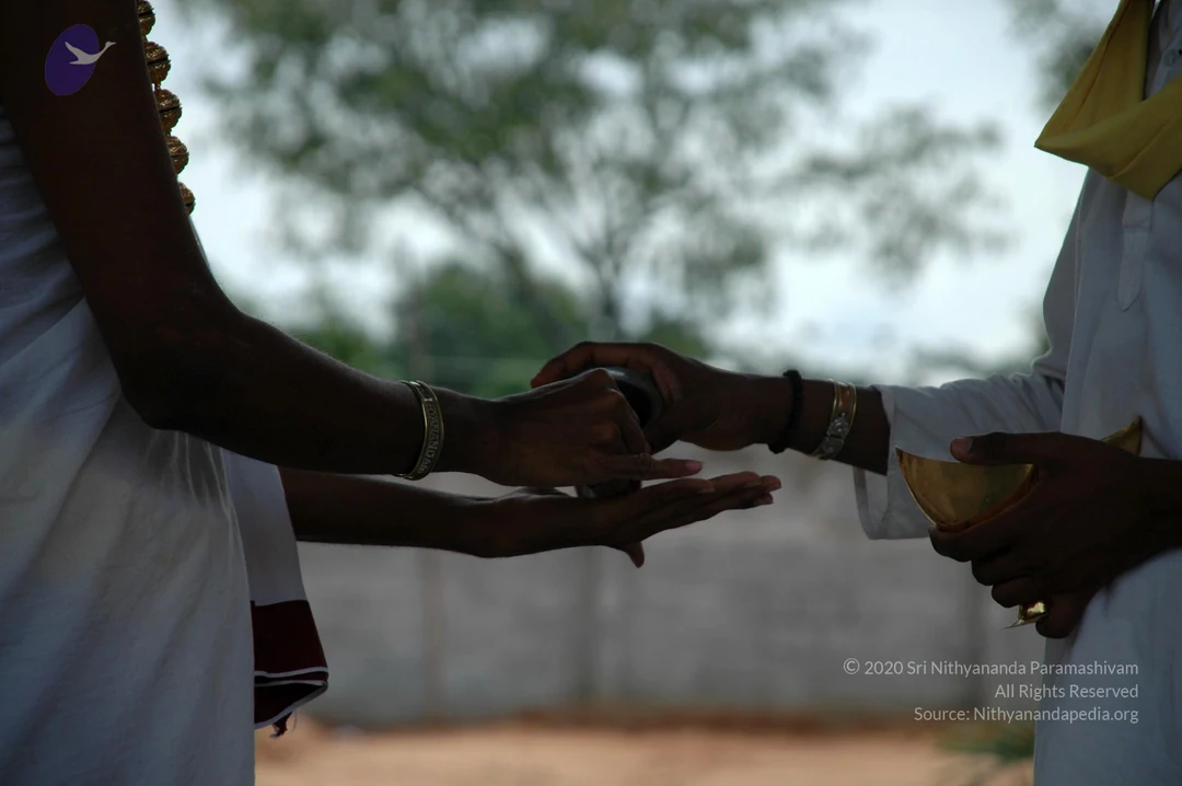 Close photograph of two people's hands and arms in a ritual exchange outdoors. On the left, a person in white clothing with a rudraksha-style bead necklace and gold bangle extends one hand to receive a small dark vessel. On the right, a person in white with a yellow cloth draped over the shoulder holds a brass bowl in the other hand while passing the vessel. A blurred tree and pale wall form the background, with dry ground at the bottom.