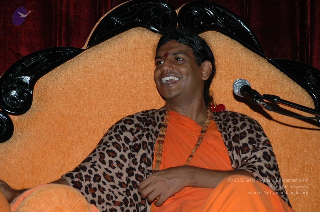 A smiling man with long dark hair and a red bindi sits on an ornate orange cushioned throne with a carved black wooden back, against a deep red velvet backdrop. He wears an orange saffron robe, a leopard-print shawl draped over his shoulders, and multiple wooden rudraksha beaded malas around his neck, with a red flower garland. A microphone on a stand is at right.