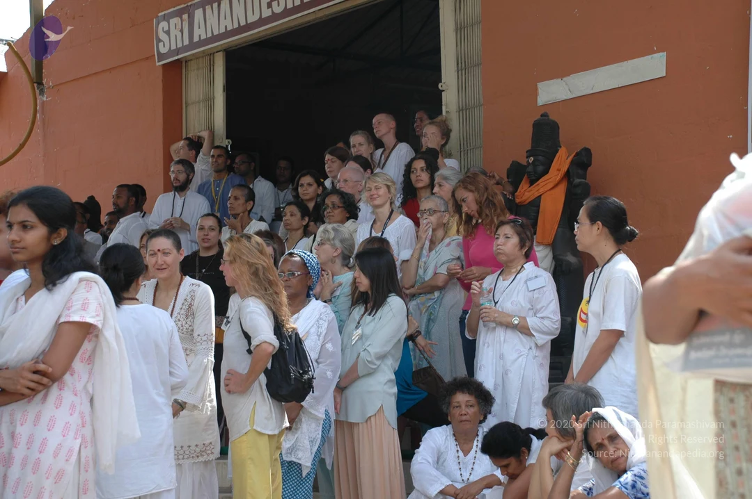 Outdoor photograph of a crowd of devotees gathered on and around the steps of an orange-walled building, beneath a sign reading "SRI ANANDESH..." above an open doorway. Many people wear white or pastel ritual clothes, some with rudraksha-style beaded necklaces and lanyards. A young woman in a white and pink patterned top stands at left, a woman in a blue headscarf stands centre, and a woman in a pink top stands near a dark stone deity murti draped with an orange cloth at right. Several women sit on the lower steps at right. The people stand and watch attentively, as at a gathering.