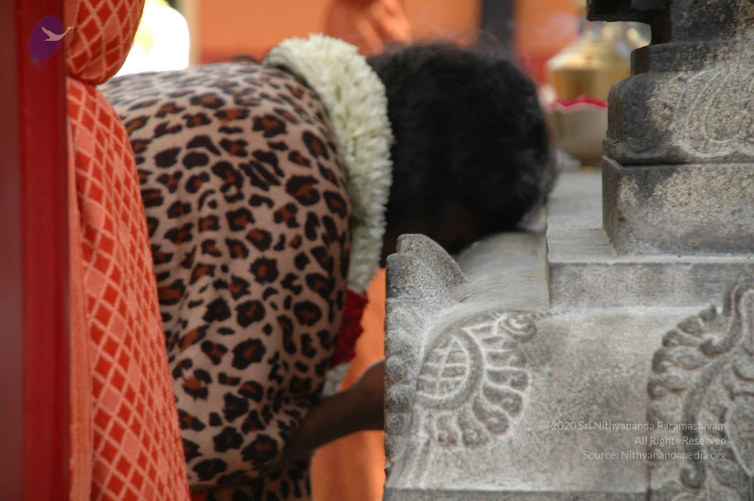 A woman in a leopard-print top and a white flower garland bends forward, head down, touching a carved grey stone pedestal with her hands. Behind her a red-and-orange patterned cloth hangs at the left edge, and a brass vessel with a pink flower sits on the stone in the background. The carved stone shows floral and scroll motifs.