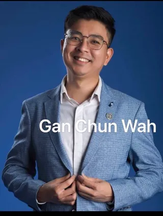 A studio portrait of a smiling man in rimless glasses and a blue textured blazer over a white open-collared shirt, with a small lapel pin, standing against a plain blue backdrop with hands holding his jacket front. A white caption reading "Gan Chun Wah" is overlaid across his chest.