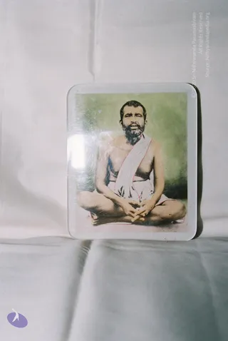 A photograph of a hand-coloured printed picture mounted behind a clear plastic or glass sheet, lying on a plain light grey cloth. The printed image shows a bearded man with short dark hair seated cross-legged in meditation posture, bare-chested, wearing a white wrap with a thin red edge draped over one shoulder, hands clasped in his lap against a soft green background. The picture is glossy and shows light reflections. The figure is a printed depiction, not a physical person present in the scene, and the printed image carries no visible caption or name.