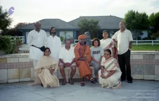 Outdoor group photograph on a paved patio in front of a suburban house with a tiled roof, white fence and green lawn. Eight people pose around a low stone wall. A central seated figure wears an orange-saffron robe, orange turban, and a rudraksha mala, holding a small green item in clasped hands. Beside them on the wall, a man in a white shirt and shorts sits with hands folded. Behind, a man in a white kurta with a black belt stands on the left, a bearded man in white stands next to him, a woman in a white sari with a red border stands at right, and a man in a white polo shirt and dark trousers stands at the far right. Two women in sarees kneel in front, one in a pale gold sari and one in a white sari with a green border. A woman in a grey DKNY t-shirt sits beside the central figure. A stone statue stands in the background.