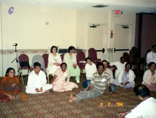 A photograph of a seated group of devotees on a patterned maroon carpet in a plain hall with cream walls, red upholstered chairs, a tripod at left and a red EXIT sign above the doors. A woman at left in an orange-and-red dupatta and patterned salwar sits smiling. A man in white kurta pyjama sits cross-legged beside a woman in a pink salwar kameez. A man in a striped grey-and-white shirt and grey trousers sits barefoot in the centre foreground, with a man in white at the lower right edge. Several women in white and pastel outfits sit behind them, including one in lavender and one in a green salwar, and a standing man in white at the far right. A date stamp reading 2 7 '04 appears in the lower right.