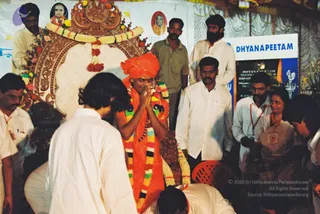 A photograph of an indoor ceremony beneath a decorated stage with a large ornate golden-brown throne-style seat draped with white cloth, flower garlands and tassels. A standing figure in an orange-red turban and orange shawl, with a multicoloured flower garland and rudraksha-style beads, holds hands folded in namaskar. Around him stand men in white shirts and dhotis, and a man in an olive shirt; a woman in a cream sari with a lanyard watches at right. In the foreground, a long-haired man in white clothes bows with his back to the camera, and another man bends low at the bottom edge. Behind them is a blue and white banner reading DHYANAPEETAM, with a black panel carrying more text, and portrait photos of robed figures are displayed on the backdrop. Several people wear lanyards.