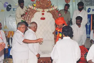 A photograph of an event stage with an ornate carved wooden throne upholstered in white quilted fabric, draped with a garland of yellow, orange and green flowers and red pom-pom tassels. A man in an orange-saffron turban and a multicoloured beaded garland stands smiling beside the throne. Two older men in white shirts and dhotis, one wearing glasses, lean in and gesture toward the throne. A bearded man in a white kurta with his back to the camera stands in the foreground. Several men in white kurtas, some wearing red lanyards, stand around the stage. Behind them hangs a white backdrop with a blue banner and a brass oil lamp. The orange-turbaned man is a person by his saffron turban and colourful garland, but the appearance is uncertain.