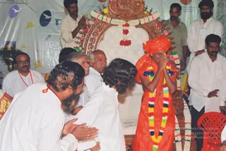 A photograph of a ceremonial stage in front of an ornately carved wooden throne draped with white and multicoloured flower garlands. A figure in a bright orange turban, orange robe and a long rainbow-coloured garland with blue and gold pendants stands at the centre with palms pressed together at the face. Several men in white kurtas, some with red neck cords, bow or stand with hands folded beside and in front of him, and a bearded man in white bends forward toward him. Behind them, men in white and khaki clothing stand near a white backdrop decorated with yellow leaf motifs, and a red plastic chair sits at the lower right. a person is the orange-robed figure with turban, garlands and the ornate ceremonial necklace.