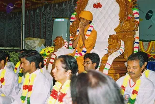 A seated figure in saffron-orange robes and a saffron turban sits on an ornate carved wooden throne flanked by two carved lion sculptures, with white quilted upholstery behind. He holds his palms pressed together in namaste. Multicoloured flower garlands in red, yellow, white and green drape the throne and the lions. In front, several people in white clothes wear floral garlands: a man on the left with his eyes lowered, a woman in a white sari with her eyes closed, and a man in white with a red lanyard on the right. Black curtains and potted plants form the backdrop.