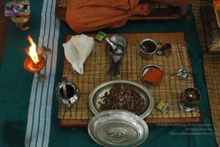 A ritual setup on a woven straw mat over a teal cloth with white stripes. A lit oil lamp with a flame sits at the left edge. Centre features a silver plate of brown seeds or grains, a white conch shell, a small silver pot with a spout, a silver ladle, a small bowl of red powder, a brass stand with incense sticks, and a silver container with a small flame. A partial figure in orange saffron cloth sits at the top edge, mostly out of frame. Matchboxes lie on the mat. This is a photograph of puja articles arranged for worship.