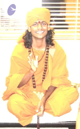 A smiling young man sits cross-legged on a light surface in front of white-slatted window blinds, raising his right hand in a blessing gesture while holding a rudraksha mala in his left. He wears a saffron-orange turban with a red tilak on the forehead, a saffron-orange robe and loose saffron trousers, and a long rudraksha mala with silver spacers hangs around his neck. Long dark wavy hair falls past his shoulders.