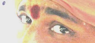 Extreme close-up photograph of a person's face showing one eye and eyebrow, with a red kumkum mark on the forehead between the brows. The skin tone is warm and the image is heavily washed out with bright highlights. A reddish-orange fabric edge is visible at the top right of the frame. No other features of the setting are visible, and the photo shows no ritual activity clearly enough to name.
