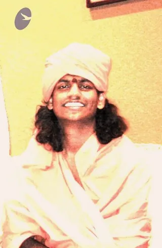 A smiling man with long dark hair sits against a pale yellow wall, wearing a cream-white turban and a cream-white shawl draped over his shoulders and arms. A red bindi-style mark is visible on his forehead. A dark framed picture hangs at the upper edge of the wall. The warm, saturated colour gives the photo a yellowed tone.