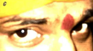 A close-up photograph of a man's face showing the eyes, eyebrows and forehead. A yellow-orange cloth edge is visible at the top of the frame, and a red kumkum mark sits on the forehead between the brows.