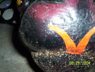 A close-up photograph of a dark, glossy, rounded object. A yellow glass-like eye or bead is visible at the top left edge, and a patterned dark surface with pale spots sits behind it. The object rests on a grey concrete floor. No people are visible. A date stamp reads 08/28/2004 in the lower right corner.