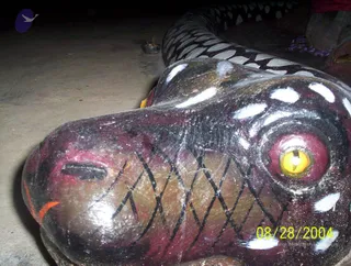 Close-up photograph of a large painted wooden serpent head lying on a concrete floor, its body coiled behind it in black and white banded scales. The head is maroon and brown with black diamond-pattern markings, white spots, a glossy yellow eye with a dark pupil, and a red-orange mark near the snout. A small clay oil lamp with a wick sits on the floor in the background, and a purple cloth is visible at the top right. No people are visible.