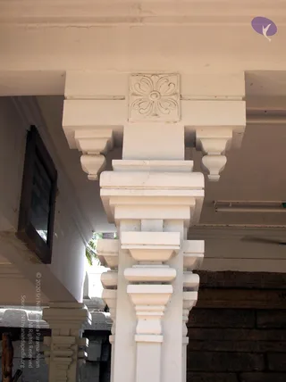 Close-up photograph of a white-painted stone pillar in a temple-style colonnade, shot looking upward. The capital has a square carved rosette panel with a stylised flower of looping petals, flanked by bracket corbels with cup-like drops. Below, the shaft has stepped, tiered mouldings and projecting ledges. A dark wooden framed board hangs on the left wall, and another pillar with similar stepped carving appears in the background. No people are visible.