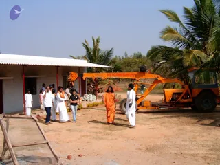 A dusty outdoor construction site beside a white single-storey building with a corrugated roof and red pillars. A bright orange crane-and-digger vehicle with a boom reading G.T. CRANE SERVICES stands on the right beside coconut palms. In the centre, a man in an orange-saffron dhoti and shawl with long dark hair walks across the sand; he has distinctive long swept-back hair and saffron clothing, and the page does not place him at this event, so his identity is unsure. Around him, several people in white dhotis and kurtas stand and talk, including a woman in a dark top and jeans and a young child in pink. A stack of white sacks and wooden planks lie on the ground.