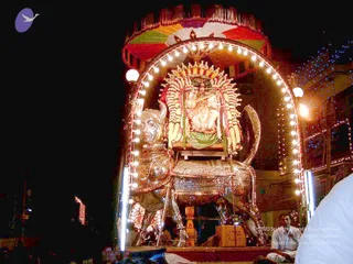 Night photograph of a festival procession vehicle lit by rows of bulbs framing an arch. A silver-gilded lion-like mount with a carved head and ornate harness carries a deity murti seated on a platform, decorated with a tall garland-covered backdrop of yellow, orange and white flower garlands and a multicoloured canopy overhead. Strings of coloured lights line nearby buildings. In the lower right, a man in a white shirt sits at the platform's edge with his hands together. The vehicle appears to be a ratha/vahana used in a temple procession.
