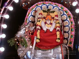 A photograph of a decorated processional deity murti seated on a silver Nandi bull vahana at night. The figure wears white dhoti and garment with an orange-saffron face cover or headpiece and a crown, and is draped in a red cloth with garlands of yellow marigolds, white flowers and maroon blossoms. A large silver arch behind it is decorated with pink, blue and green bead-like ornaments and white flowers. The silver bull has green leaves in its mouth and a silver saddle with floral engraving. String lights with bulbs frame the scene.