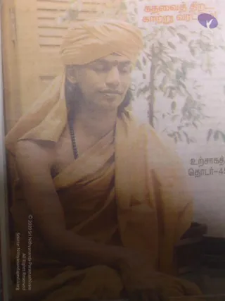 A printed photograph, seemingly from a Tamil magazine page, shows a seated young man in a saffron-orange turban and matching saffron shawl draped over his shoulders and torso. He has a red tilak mark on his forehead, a dark bead mala around his neck, and his eyes are lowered as he sits with hands resting in his lap. Behind him a window with slatted blinds and a leafy tree are visible. Tamil headline text and a purple logo sit at the top right. Probable Swamiji cues: saffron turban and shawl with a bead mala; the figure is shown as a printed depiction on the page.
