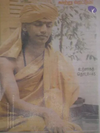 A printed newspaper or magazine page shows a seated man in an orange-saffron turban and orange-saffron robes draped over his shoulders, with eyes lowered and hands resting in his lap. A red kumkum mark is visible on his forehead and a dark beaded mala hangs at his chest. Leafy branches and a pale background appear behind him, and a wooden structure is at the lower right. A large Tamil headline in orange text runs across the top, and a Tamil label sits on the right side.