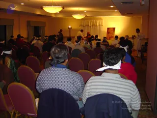 A photograph of a meditation or satsang hall with rows of maroon upholstered chairs facing a small stage at the far end. Many seated attendees, mostly seen from behind, wear white cloth blindfolds tied around their heads, and several wear plain shirts and one a red garment. On the stage, two figures in white clothing stand near a red-robed figure, with a camera operator and an attendant in white at the right edge. Warm ceiling lights and a tall floor lamp light the room, and a large portrait of a bearded saffron-robed figure hangs beside a draped white curtain on the back wall.
