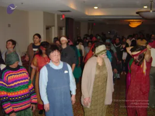 Photograph of a crowded indoor hall with beige carpet and recessed ceiling lights, where a large group of participants stands and moves in a meditative exercise. Many have blindfolds or eye coverings tied with fabric bands, including a woman in a red outfit on the right holding her blindfold up and a woman in a khaki-and-cream dress in the centre. A woman in a bright striped knit sweater with a green head wrap stands at left, and a woman in a blue denim pinafore over a light blue shirt stands in front. Others in salwar kameez and headscarves fill the background, with no clearly identifiable a person visible.