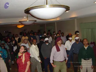 A large indoor hall with a carpeted floor and hanging white bowl-shaped ceiling lights, where a crowd of participants stands with white and maroon blindfolds or cloths over their eyes. In the foreground a woman in a red and orange patterned top with a beige scarf stands with her eyes closed, next to a man in a white shirt and grey trousers, a man in a maroon head cloth over a checked shirt, and a man in a green sweater with a white collared shirt. Behind them many other people in mixed everyday clothing stand in a group. No identifiable a person figure is visible.