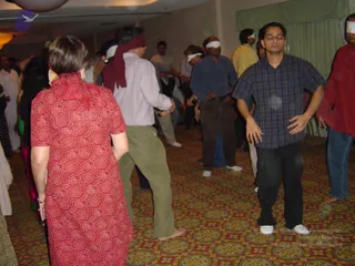 A photograph inside a carpeted hotel-style hall during a group activity. Several participants stand in a loose crowd with white or red cloth blindfolds over their eyes. In the foreground, a woman with short brown hair wears a red patterned kurta and a red scarf over her head, seen from behind. A man in a white-and-purple checked shirt and olive trousers, with a maroon cloth tied over his eyes, stands near her. To the right, a man in a dark plaid shirt and black trousers stands with eyes closed and hands at his hips. Another man in a red shirt stands at the right edge. Behind them, others in blue, yellow and white clothing stand on a red and gold patterned carpet beneath a white ceiling light and green curtains.