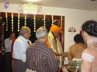 Indoor photo at a temple-style room with a green EXIT sign and yellow paper garland streamers hanging from the doorway. A man in an orange turban and gold-brocade shawl over orange clothing stands center, smiling and reaching toward a bare-backed man with white ash markings, who sits with his back to the camera. A man in a blue shirt and a man in a plaid shirt stand in the foreground, another man in a light shirt watches at left, and a bare-chested man with white markings and a gold chain holds a metal tray at right. A wall calendar and a framed picture are on the right wall.