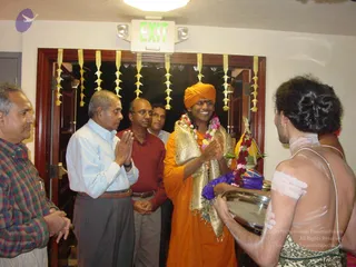 A photograph of an indoor doorway with yellow paper garlands and a green EXIT sign above. A man in a saffron-orange robe and orange turban, wearing a gold-bordered shawl and floral garlands, stands centre with palms pressed together. To the left, four men in shirts and trousers, one in a plaid shirt, press their palms in greeting. On the right, a bare-chested priest with white ash markings on his shoulders and back holds a silver tray with a flower-decorated item. The page excerpt mentions a pada puja, but the visible evidence shows a greeting with a tray offering.